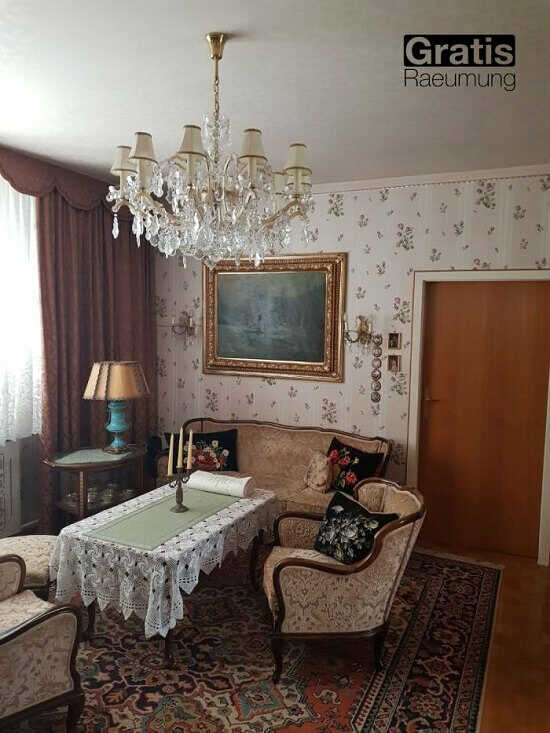Ein traditionelles Wohnzimmer oder eine Lounge. 2 Einzel- und 1 3-teilige Sitzgruppe. Ein alter, antiker Kronleuchter, Blumentapete, ein alter Lampenschirm, ein großes Gemälde und ein alter Teppich.